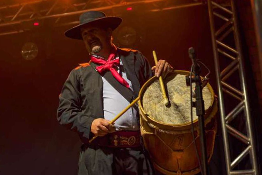 Joacir tocando bombo no palco, de chapéu e lenço vermelho